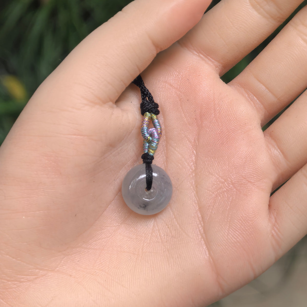 Small light gray jadeite Peace Pendant resting in an open palm, showing its compact size and black cord with a multicolored decorative knot.