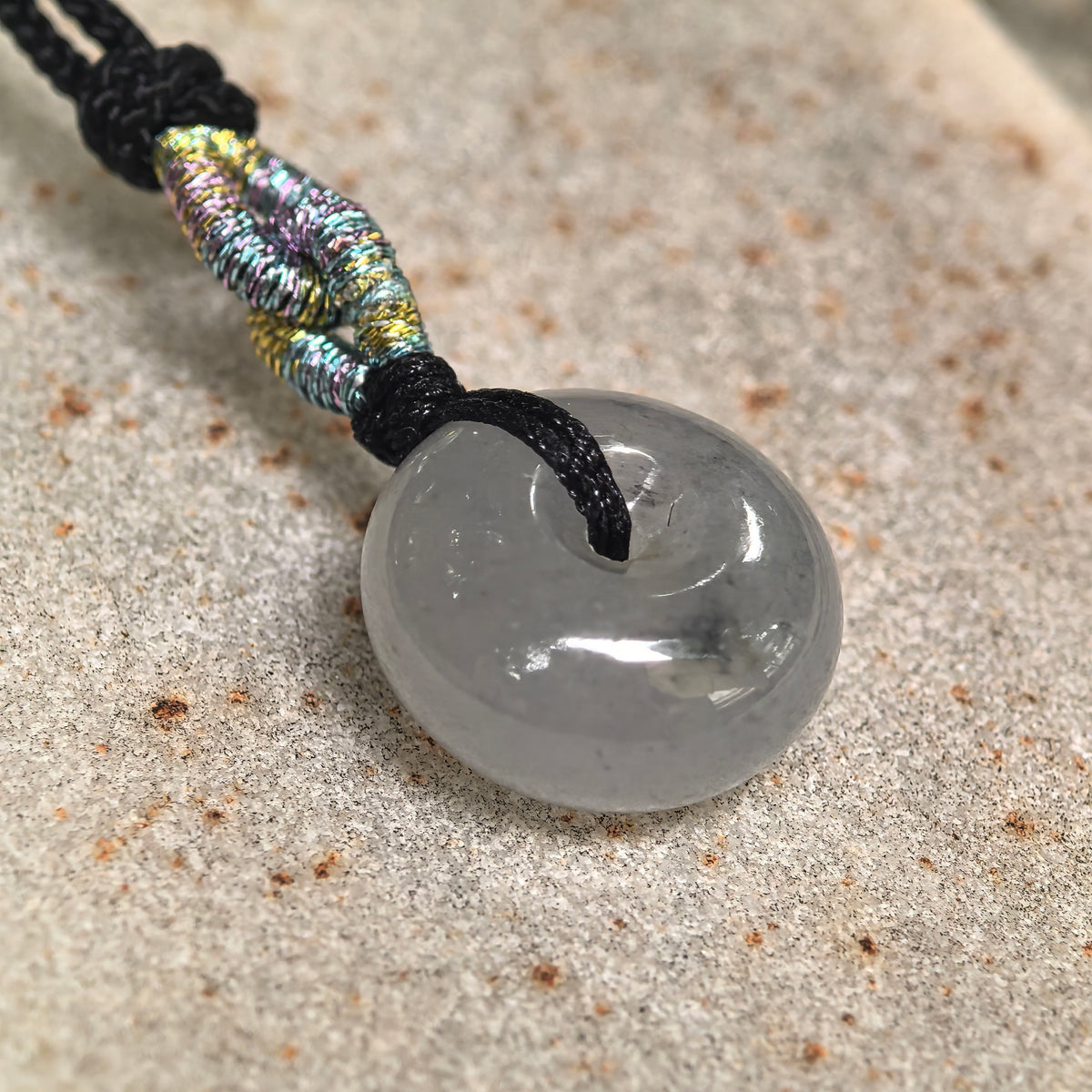 Angled close-up of a light gray jadeite Peace Pendant on stone, showing its thick, rounded profile, polished surface and ink-black floating pattern.