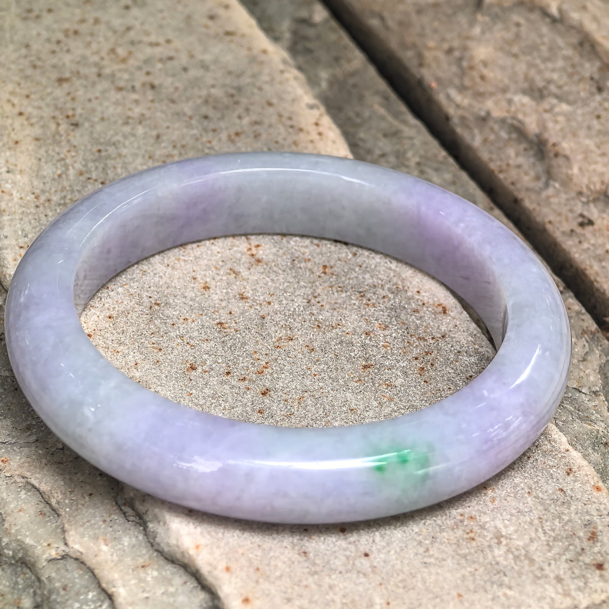 Angled view of a glutinous lavender jadeite D-shape bangle on textured stone, showing whitish intervals and a bright green accent along the front.
