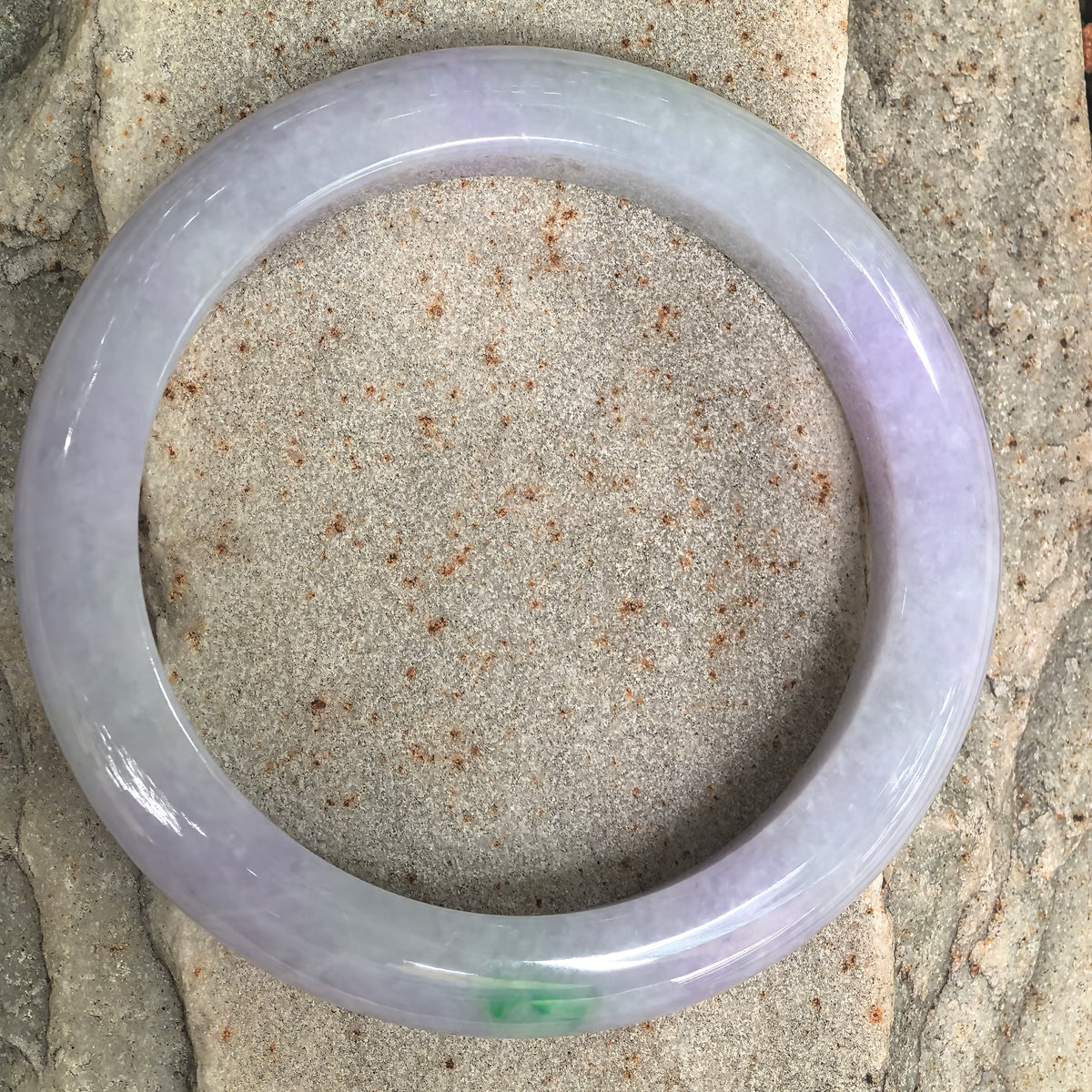 Overhead view of a lavender jadeite D-shape bangle on stone, showing its circular outline, alternating purple and whitish sections, and a green accent at the bottom.