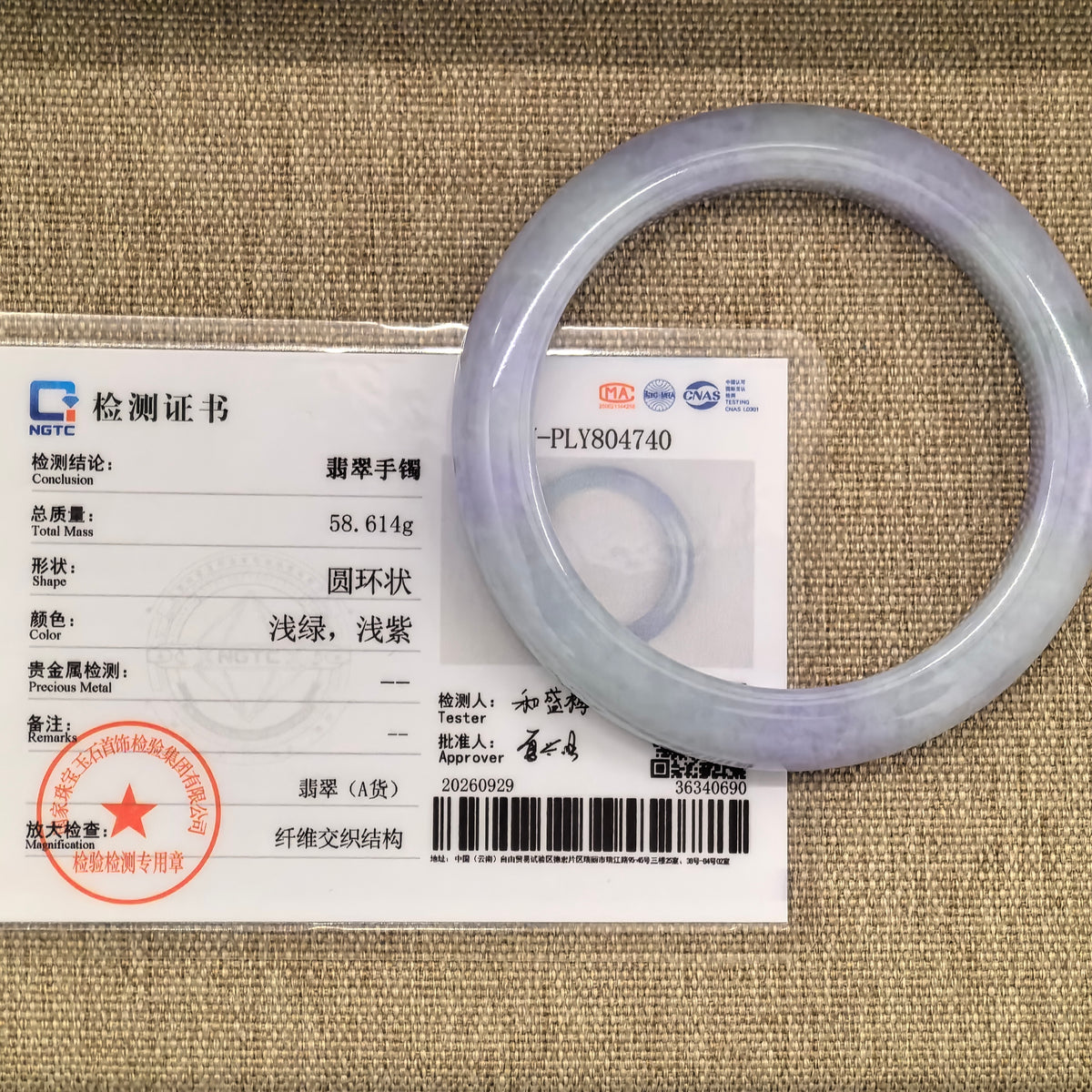 Pale lavender jadeite D-shape bangle displayed over its matching NGTC certificate on a woven background.
