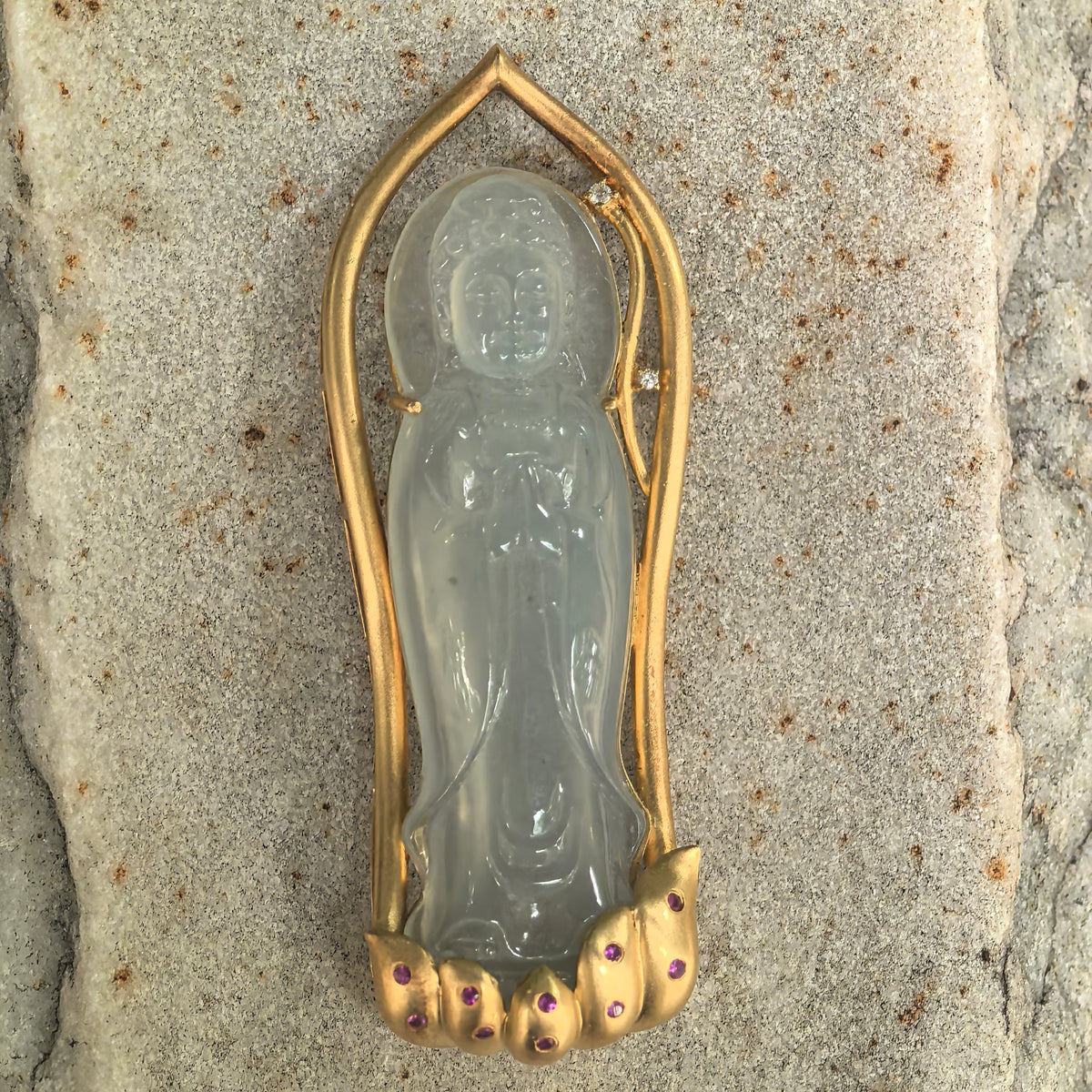 Full front view of a greenish white jadeite Guanyin pendant resting on stone, showing the elongated gold frame and decorative petal-shaped base.