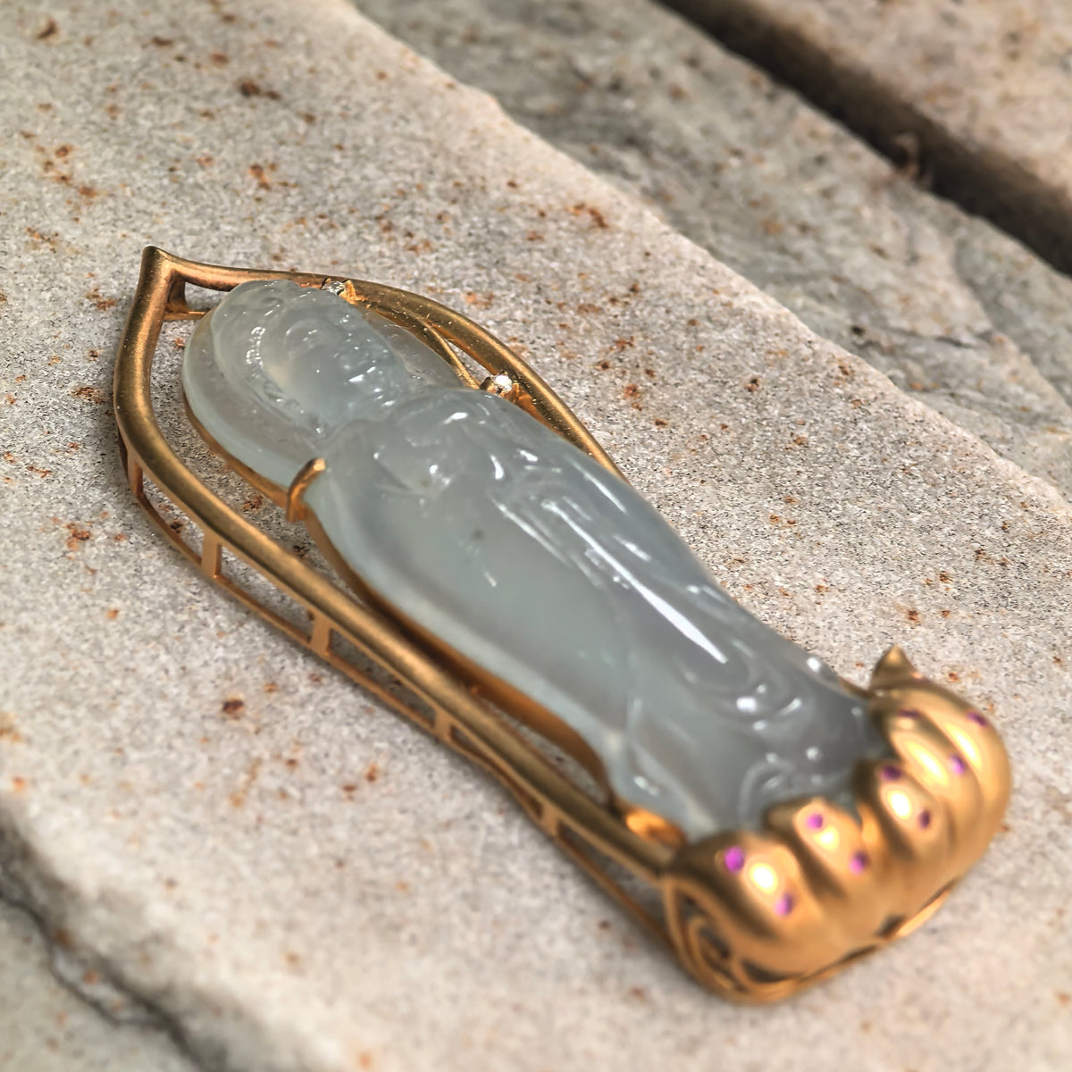 Angled close view of a greenish white jadeite Guanyin pendant on textured stone, showing the carved figure and raised 18K gold frame with a petal-shaped base.