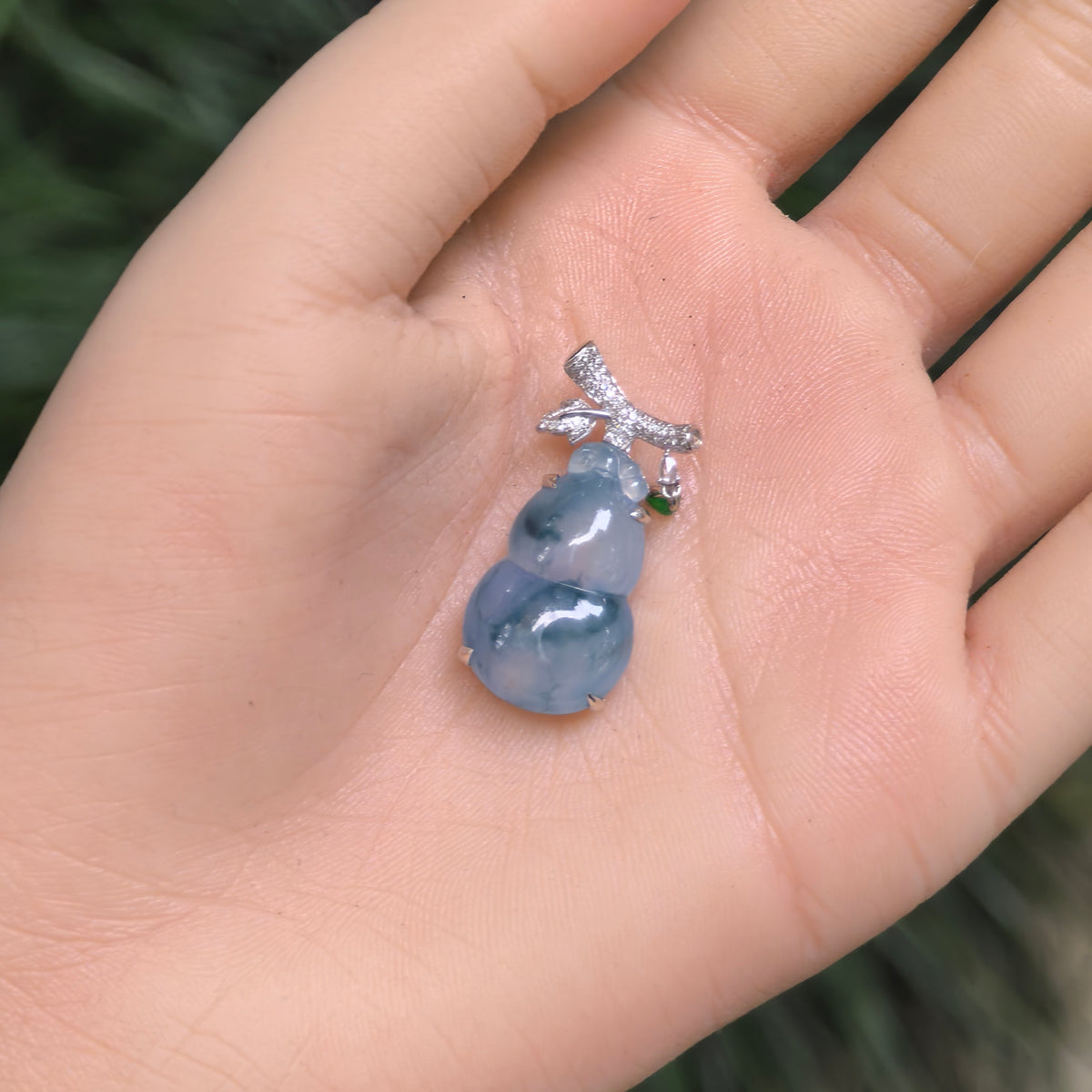 Blue-lavender jadeite gourd pendant held in the palm, showing deep green floating patterns, an ornate 18K gold setting and a small hanging green accent.
