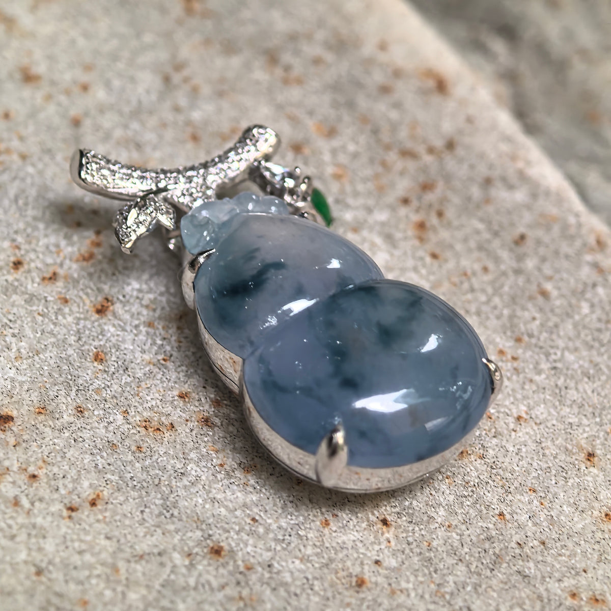 Angled close view of an icy blue-lavender jadeite gourd pendant on stone, showing its rounded contours, deep green patterns and diamond-set branch and leaf detail.