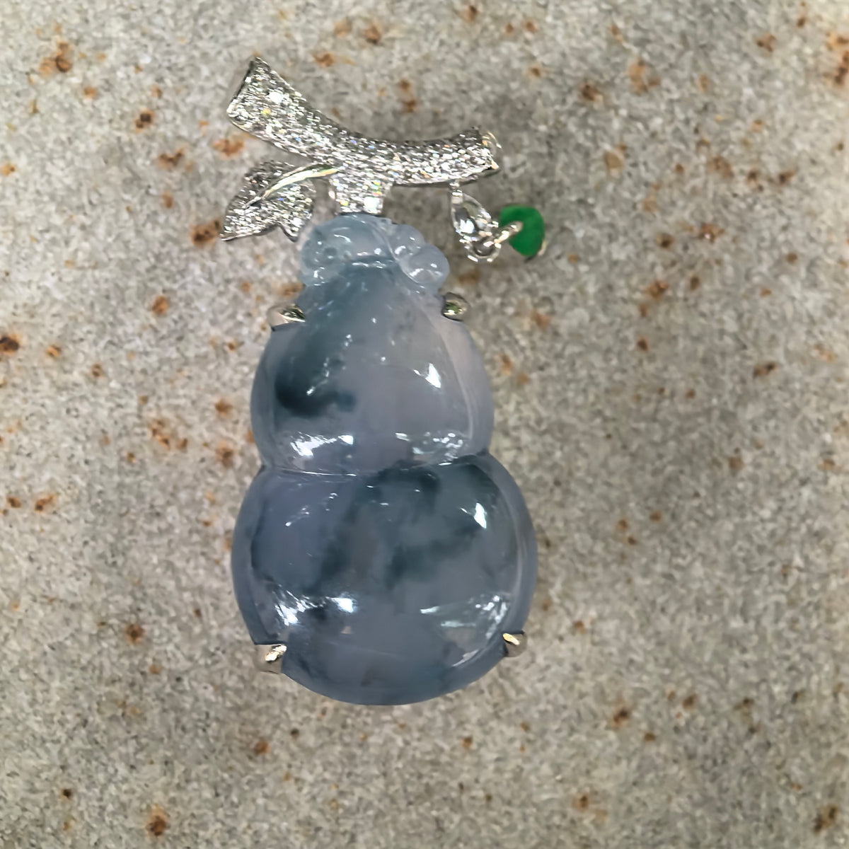 Front view of a blue-lavender jadeite gourd pendant resting on textured stone, showing the full gourd carving and decorative 18K gold setting.