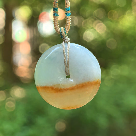 Icy glutinous white and yellow jadeite Peace Pendant hanging against a blurred green background, showing a golden-brown color band and beige cord with teal thread details.