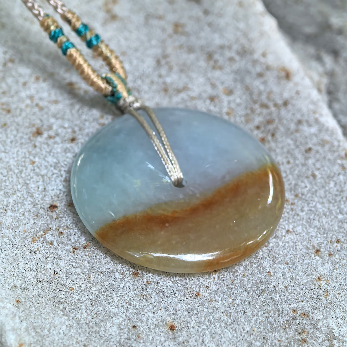 Angled close-up of the white and yellow jadeite Peace Pendant on textured stone, highlighting its polished surface, rounded edges and golden-brown band.