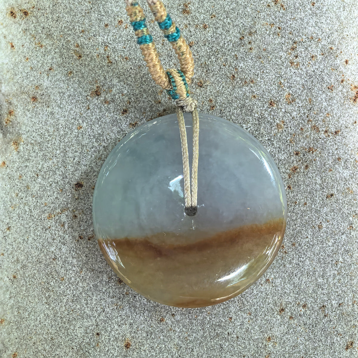Overhead view of the white and yellow jadeite Peace Pendant on stone, showing the uncarved circular face and decorative beige and teal cord.