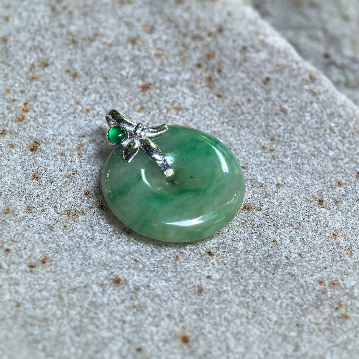 Angled view of the green jadeite Peace Pendant on textured stone, highlighting its rounded surface, floating green patterns and small green accent stone.
