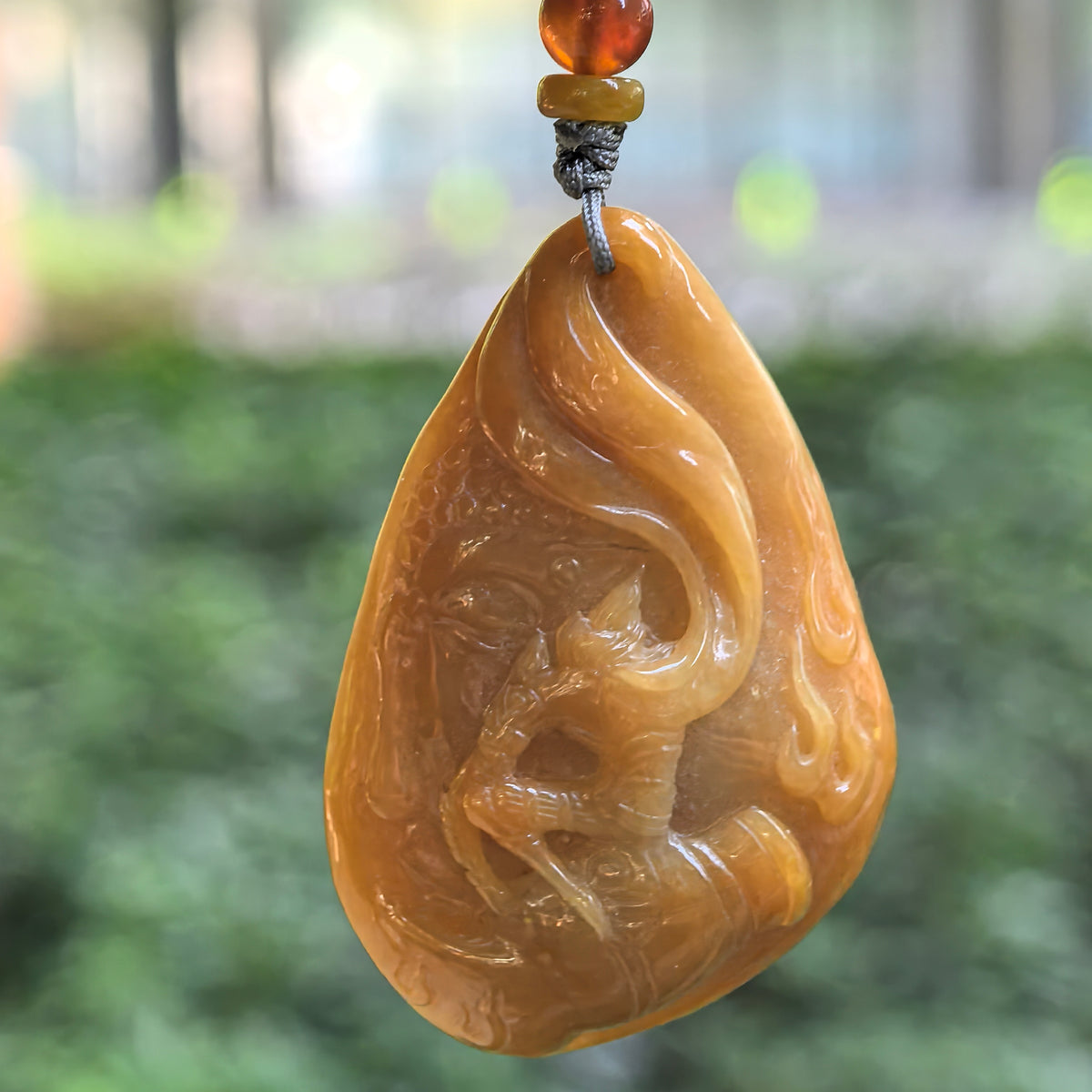 Yellow jadeite Return of the Monkey King pendant hanging against a blurred green background, with polished relief details and colored beads above the pendant.