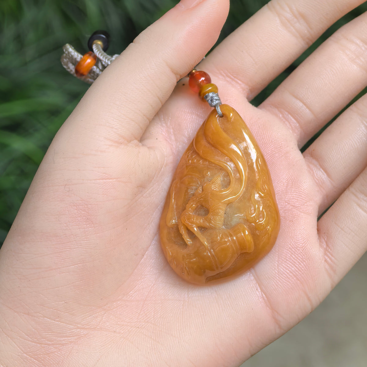 Glutinous yellow jadeite Return of the Monkey King pendant resting in a palm, showing its teardrop outline, relief carving and gray cord with colored accent beads.