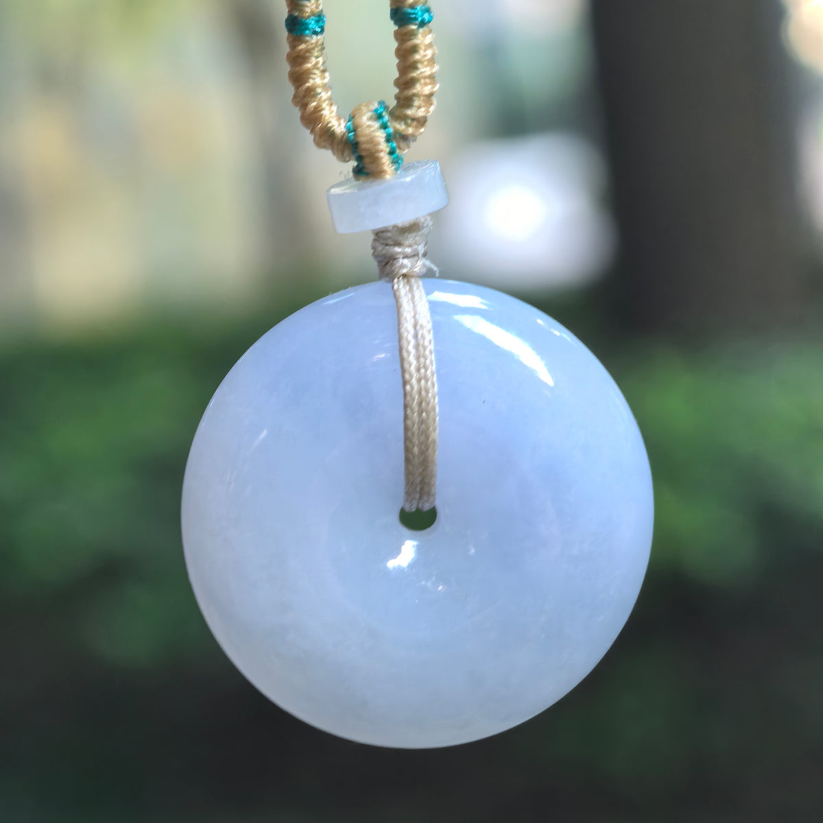 Blue-lavender jadeite Peace Pendant hanging from a braided cord with beige and teal thread accents and a pale spacer bead.