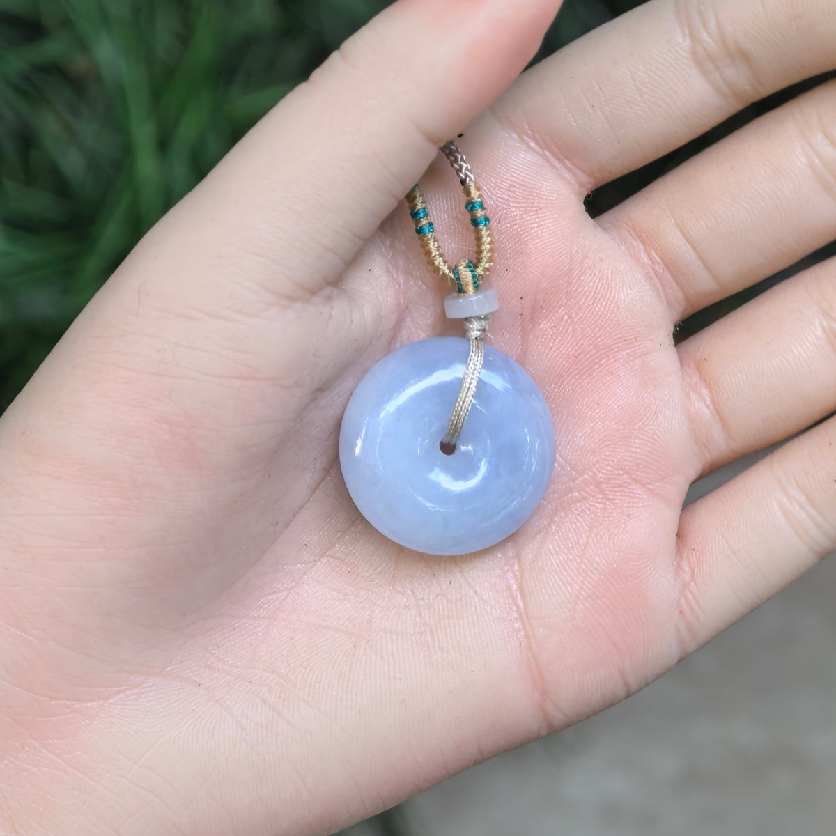 Blue-lavender jadeite Peace Pendant resting in a palm, with the braided cord and pale spacer bead visible above the circular pendant.