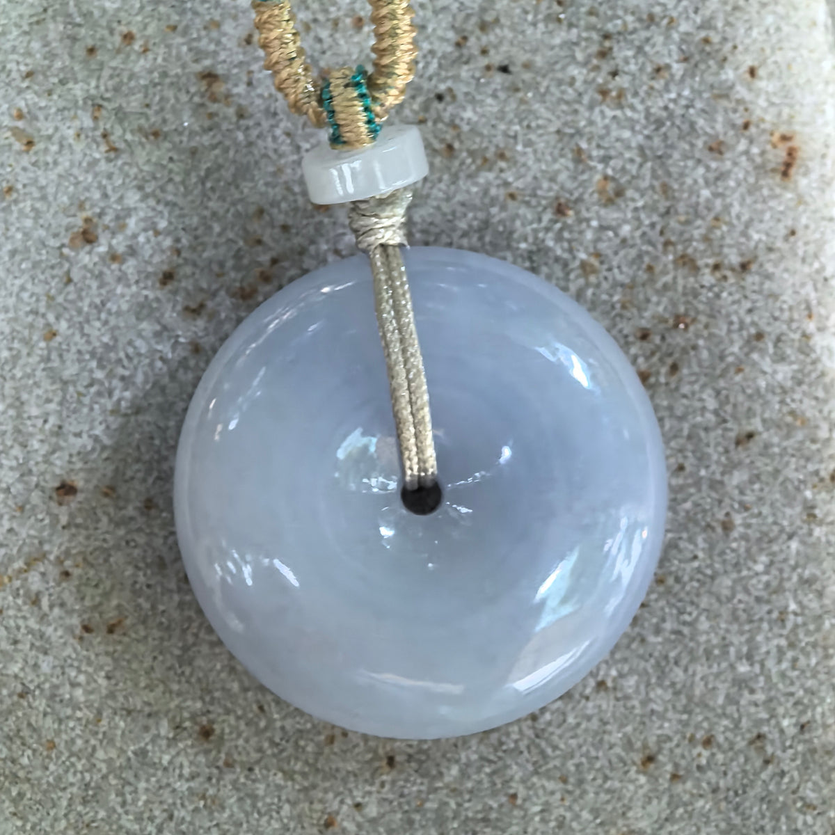 Front close-up of the blue-lavender jadeite Peace Pendant on stone, showing its soft color, smooth rounded surface, and small central opening.