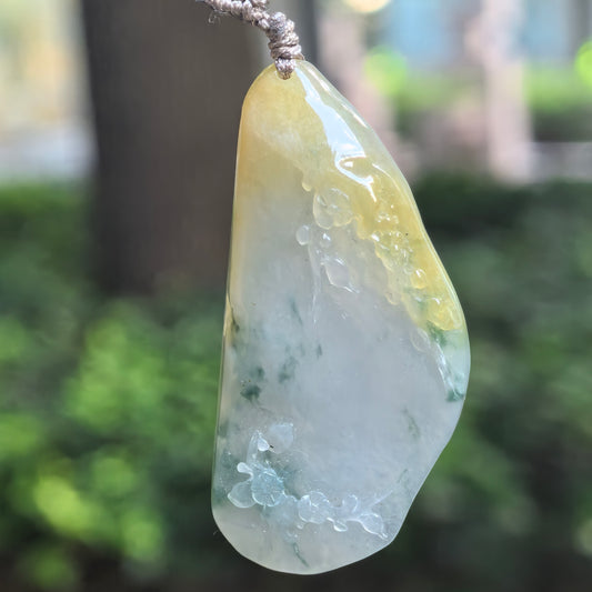 Yellow and green jadeite freeform floral pendant hanging from a gray cord, showing its translucent pale center and carved flowers along the edges.
