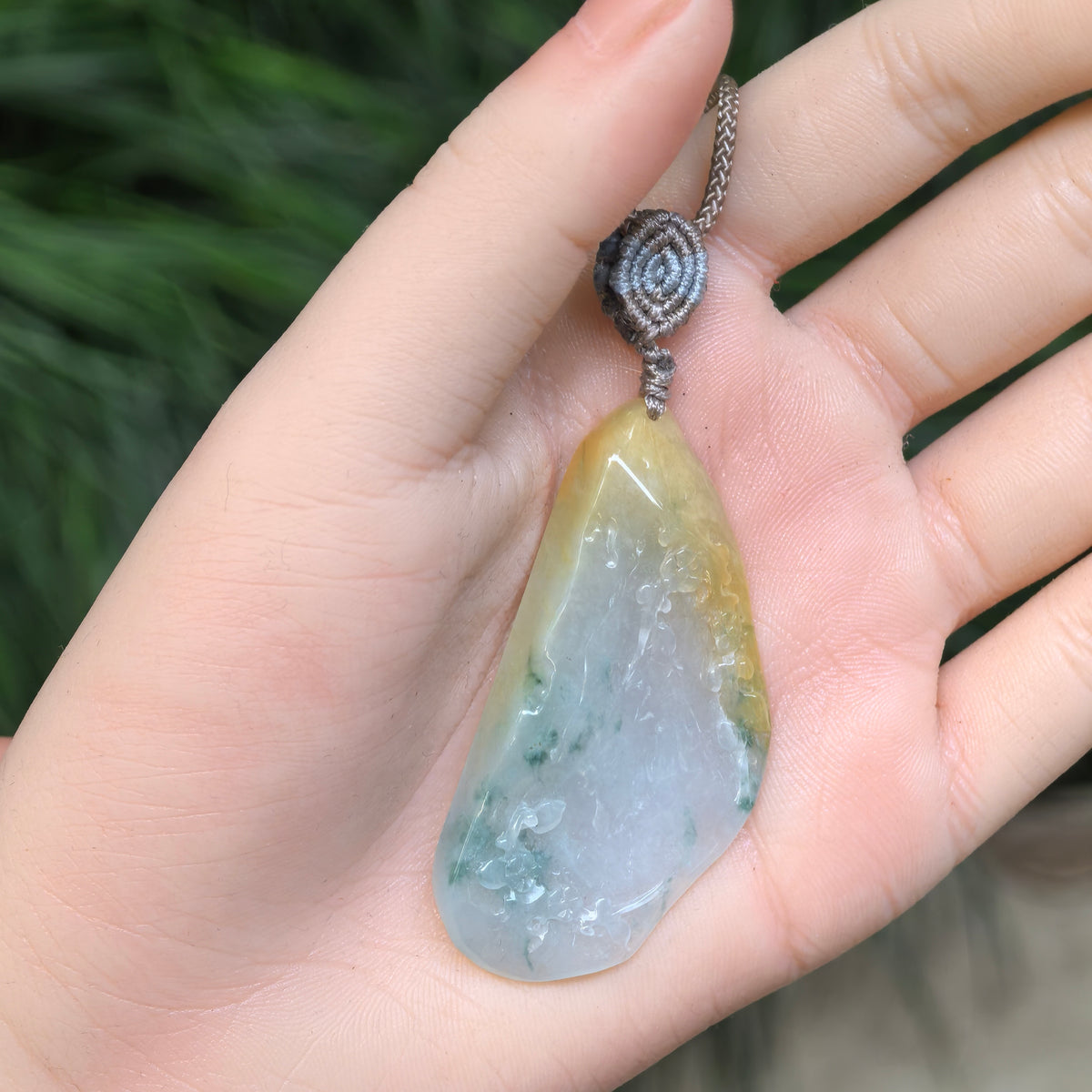 Yellow and green jadeite floral pendant resting in a palm, showing its elongated asymmetrical shape and gray cord with a round woven knot.