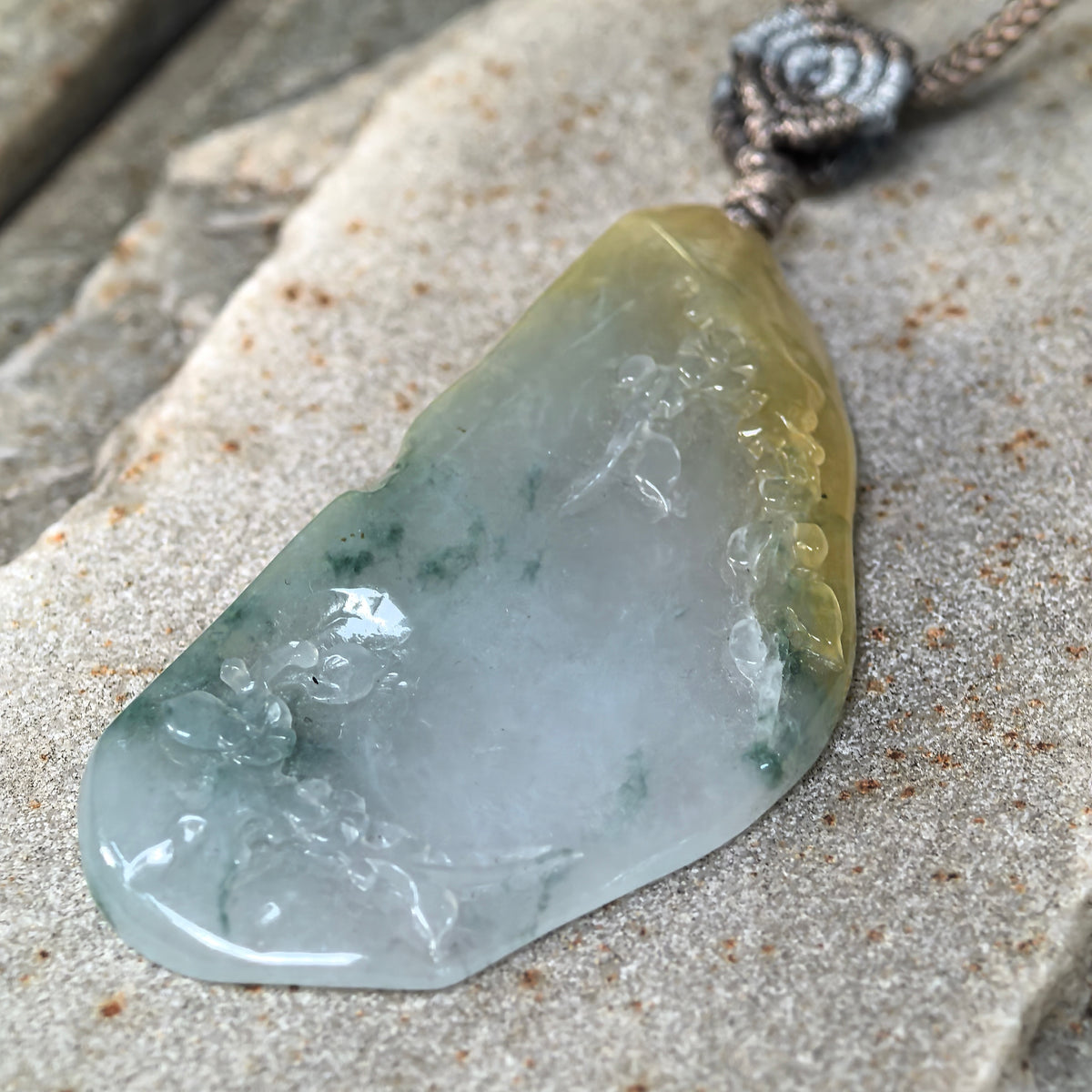 Angled close-up of the freeform jadeite floral pendant on stone, showing green patches, a yellow upper section, and the polished carved surface.