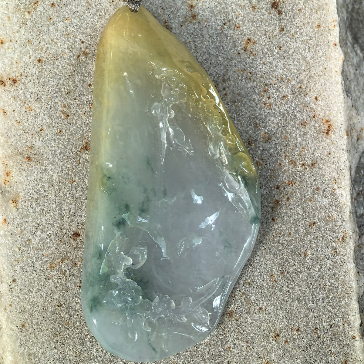 Front close-up of the freeform jadeite pendant on stone, showing yellow and green color variations and floral relief around a broad polished center.