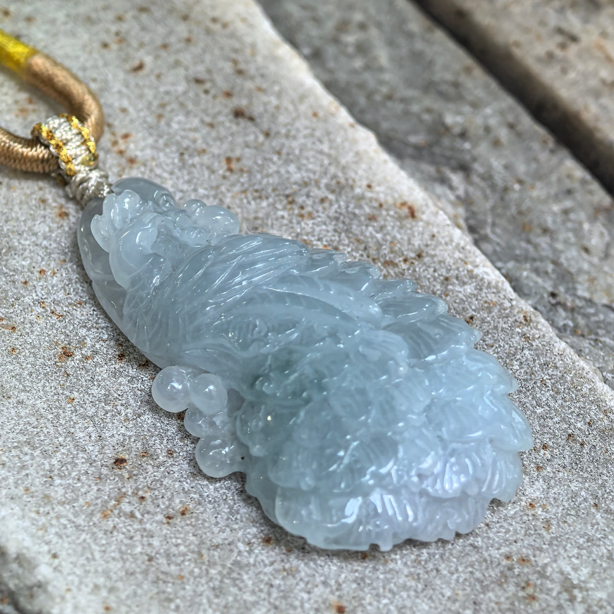 Angled close-up of a blue jadeite Phoenix Pendant on a textured stone surface, highlighting its raised carving, rounded contours and thickness.