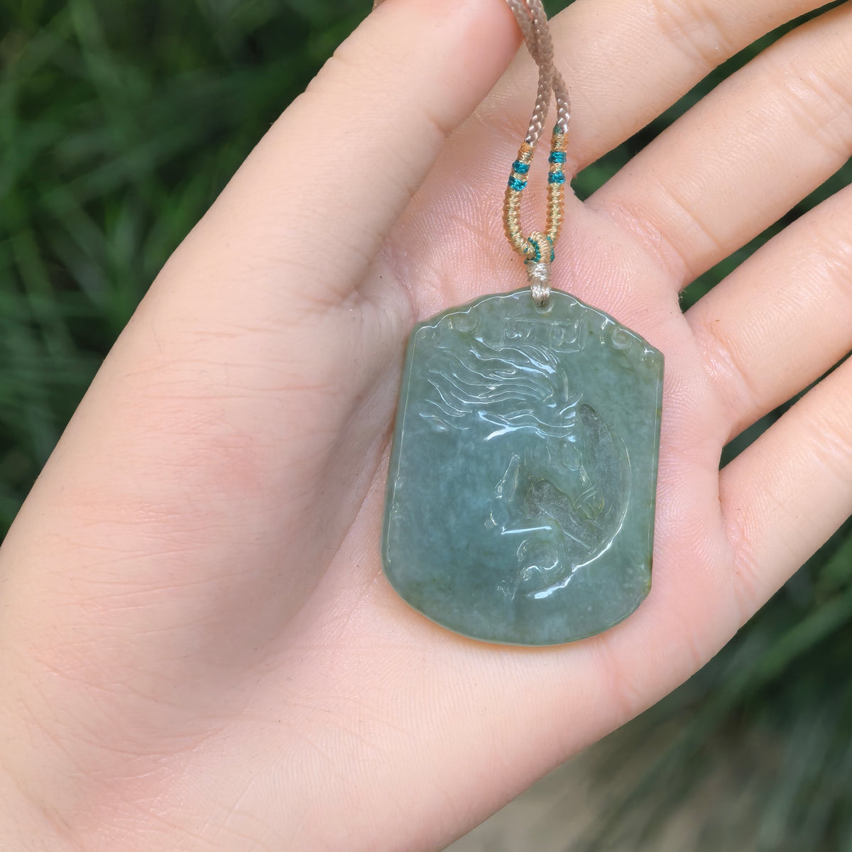 Gray-green jadeite Horse Plaque Pendant resting on a palm, showing its shaped top, rounded lower corners and beige cord with teal thread accents.