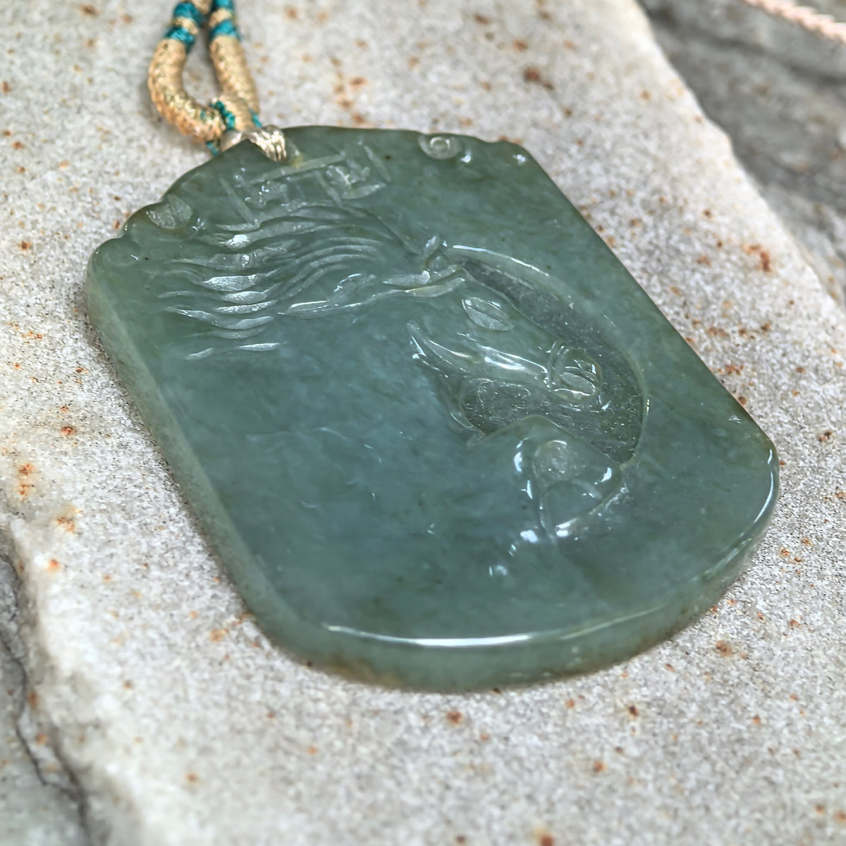 Angled close-up of a gray-green jadeite Horse Plaque Pendant on stone, highlighting the horse head relief, carved mane and polished edge.