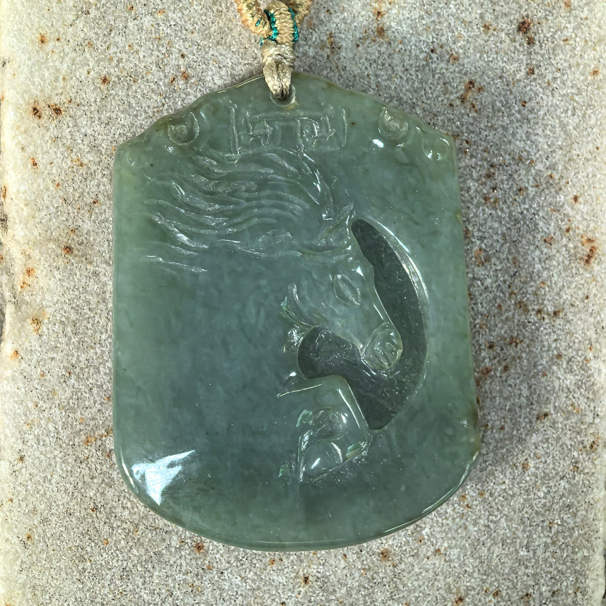 Front close-up of a gray-green jadeite Horse Plaque Pendant on a textured stone surface, showing the horse’s eye, muzzle and sweeping mane.