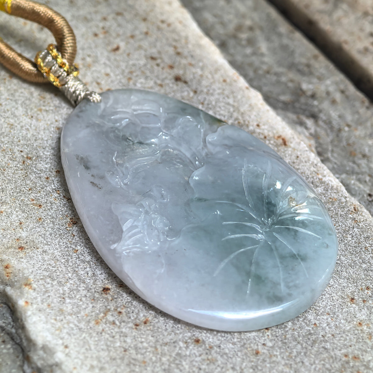 Angled close-up of a blue jadeite Floral Plaque Pendant on a textured stone surface, highlighting the relief carving, polished edge and thickness.