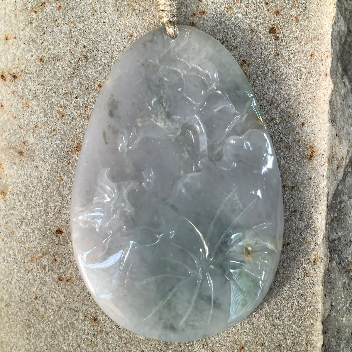 Front close-up of a blue jadeite Floral Plaque Pendant on stone, showing carved flowers, sweeping leaves and darker color patches.