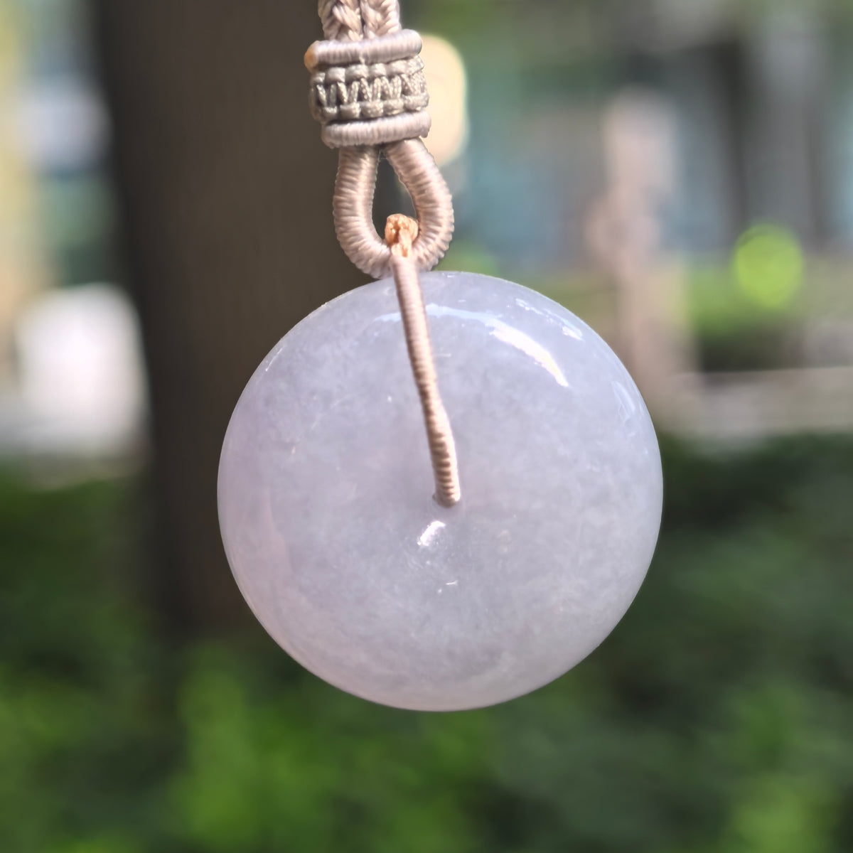 Lavender glutinous jadeite Peace Pendant hanging from a gray braided cord against a blurred green background, showing its polished circular face.