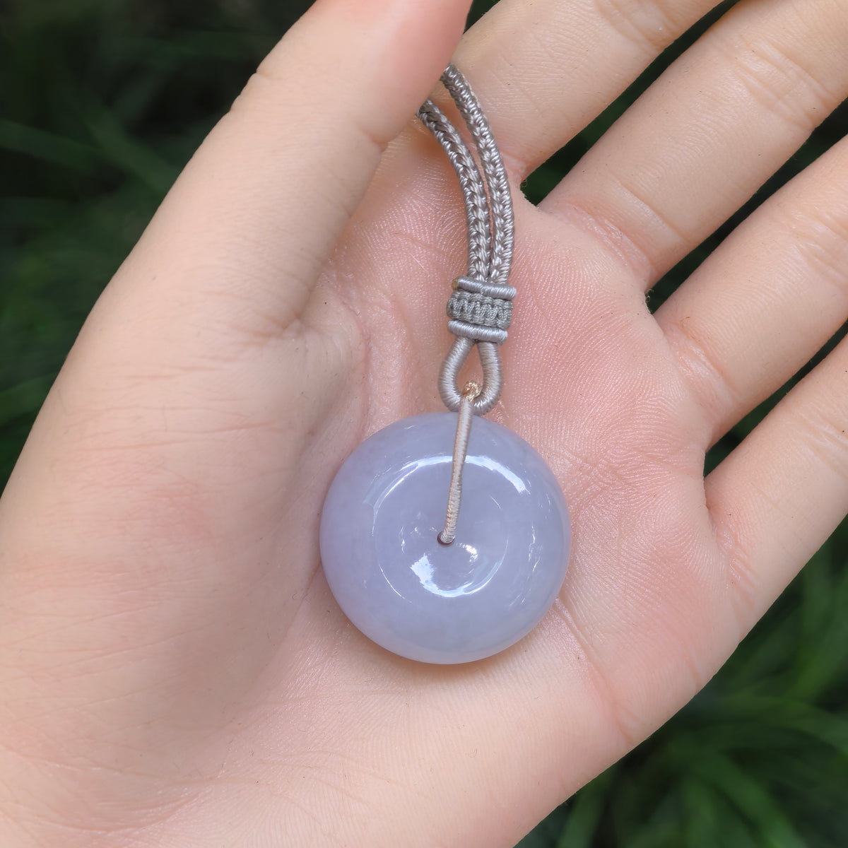 Lavender jadeite Peace Pendant resting on a palm, showing its rounded shape, small central opening and decorative gray cord knots.