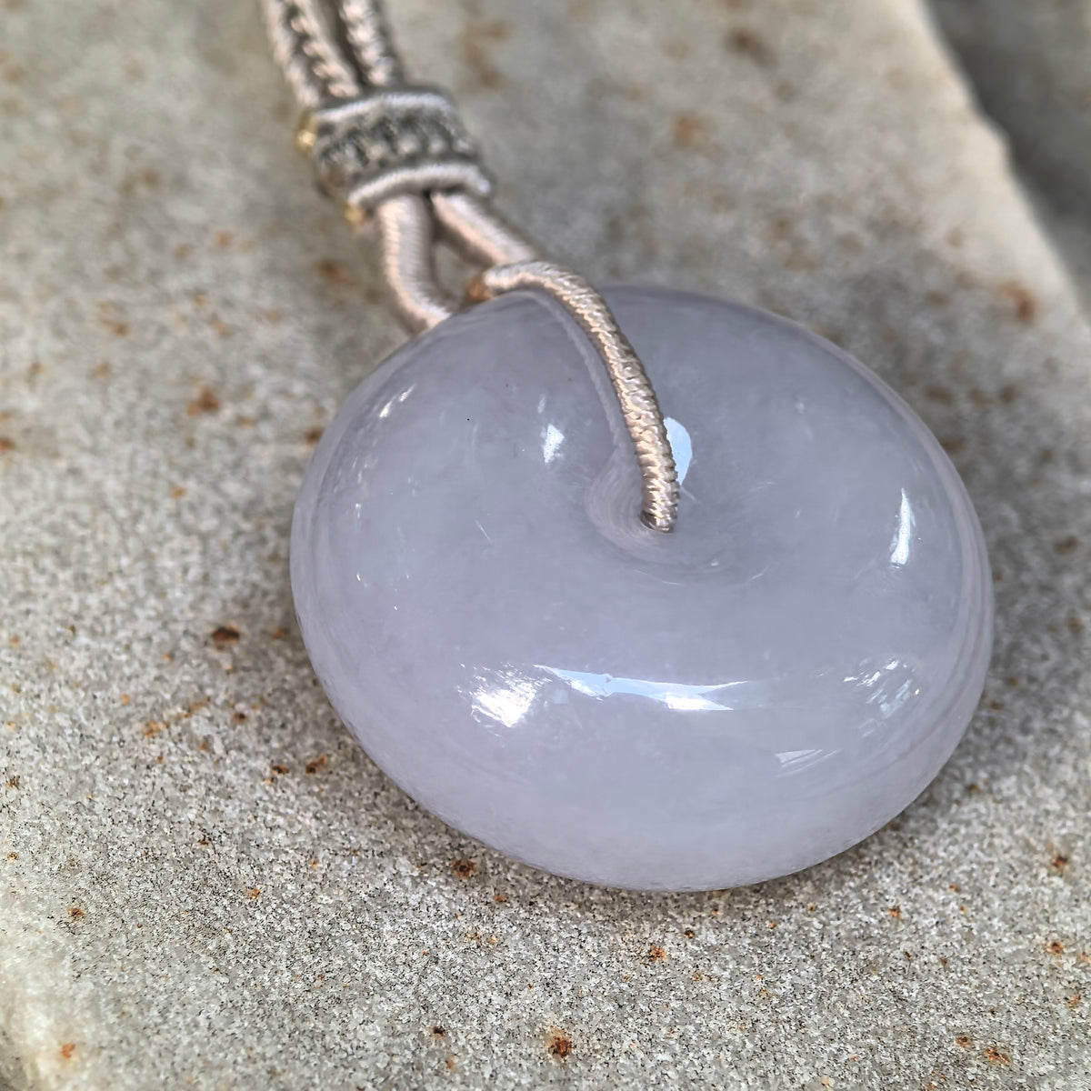 Angled close-up of a lavender jadeite Peace Pendant on stone, highlighting its substantial thickness, rounded edges and polished surface.