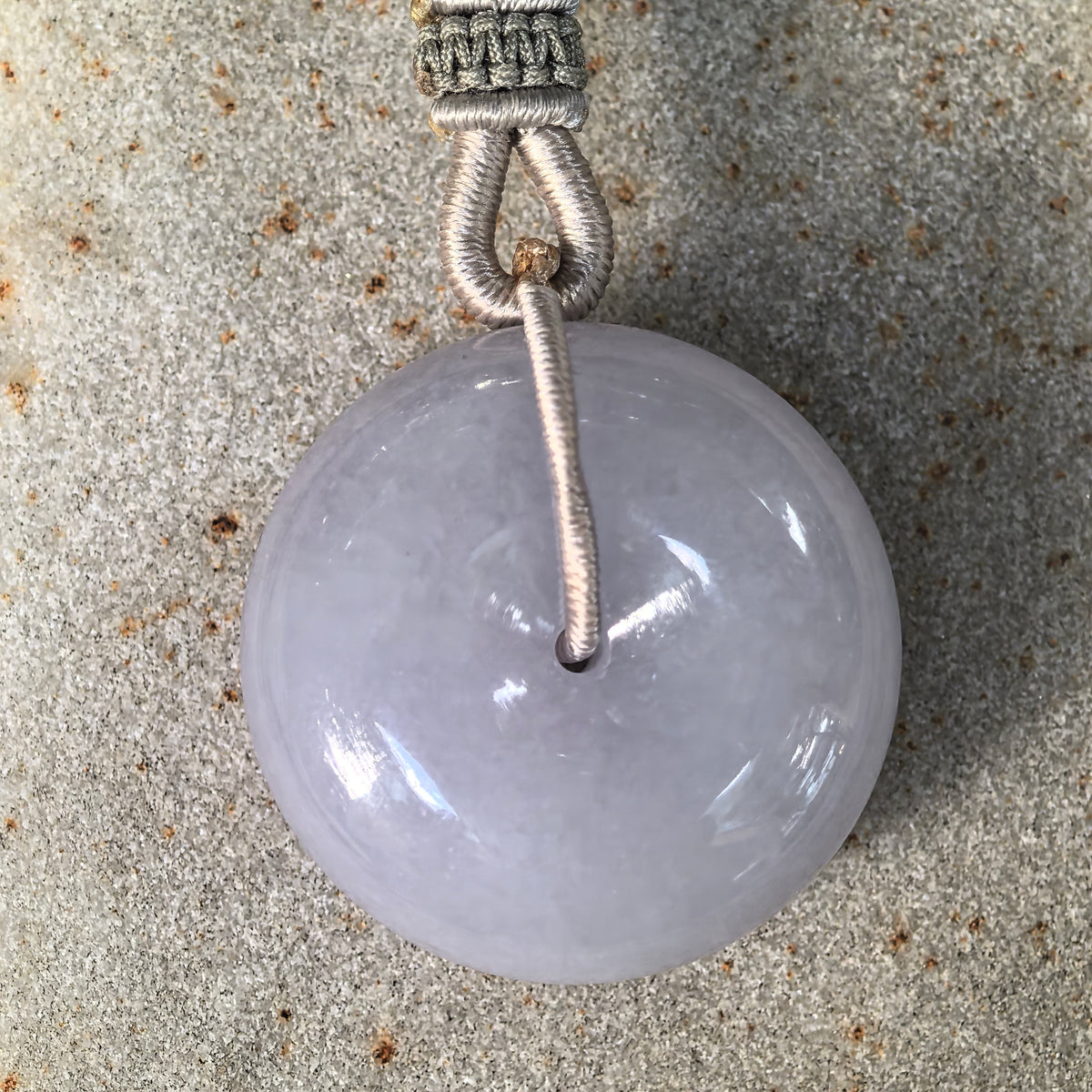 Front close-up of a lavender jadeite Peace Pendant on a textured stone surface, showing its domed face and cord threaded through the central opening.