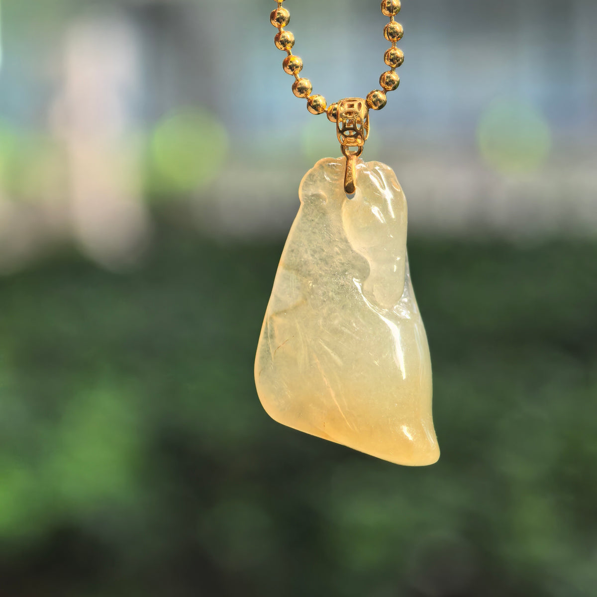 Icy glutinous yellow jadeite Horse Head Pendant with a patterned 18K gold bail, hanging from a gold-colored bead-style chain against a blurred green background.