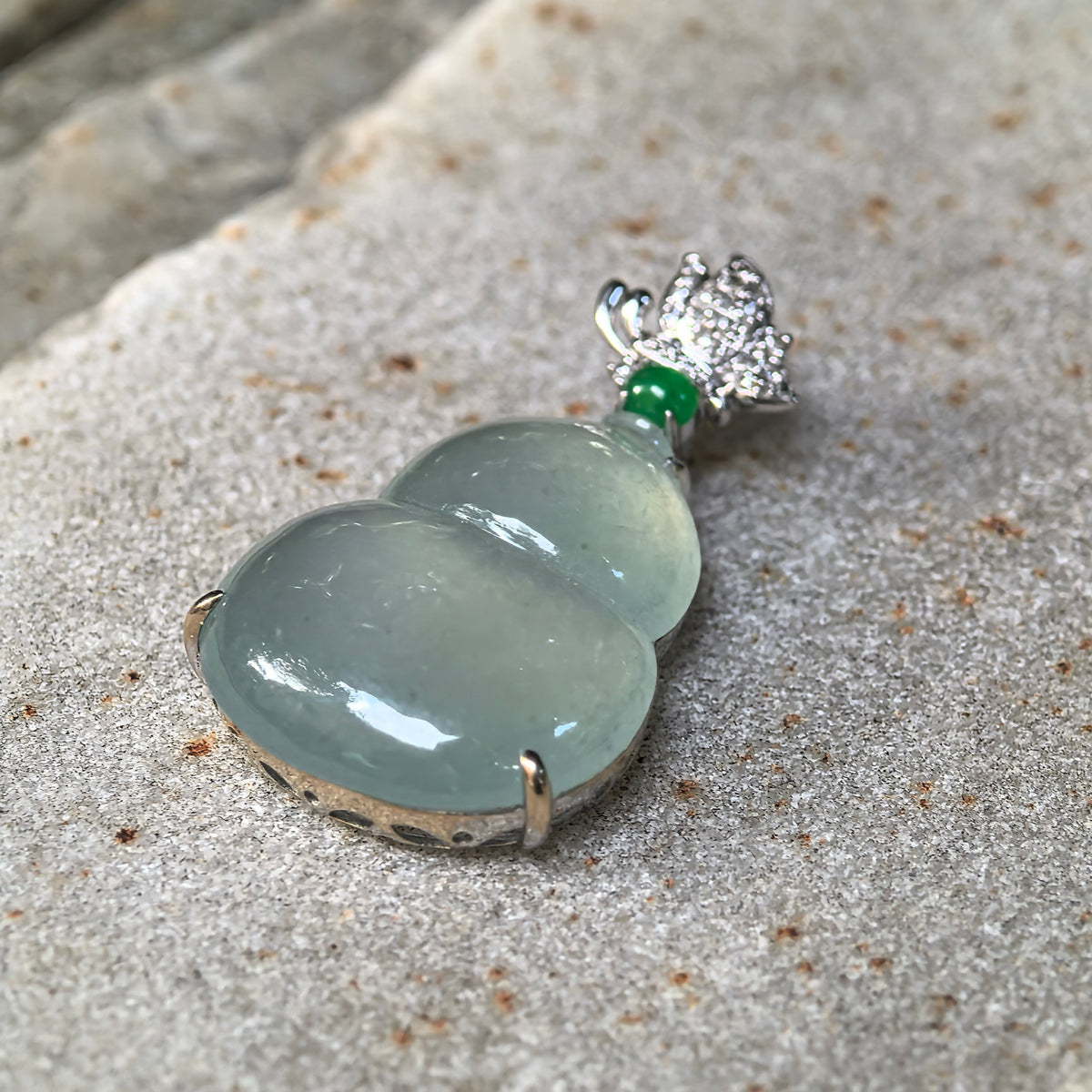 Angled view of the light green jadeite Gourd Pendant on a stone surface, highlighting its domed contours, gold prongs and the side of the setting.