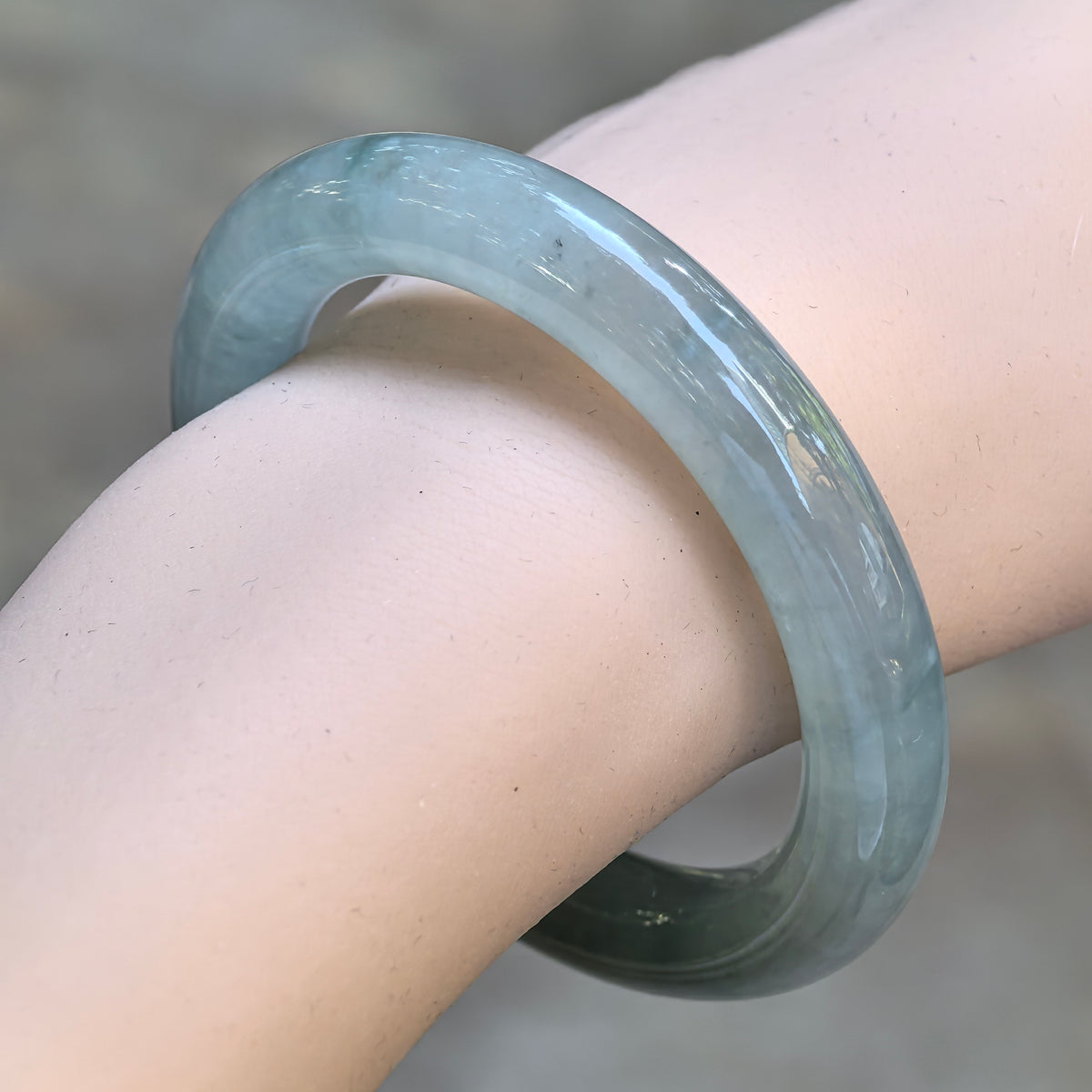 Angled close-up of a light green round-bar jadeite bangle worn on the wrist, showing its subtle blue undertone and polished rounded profile.