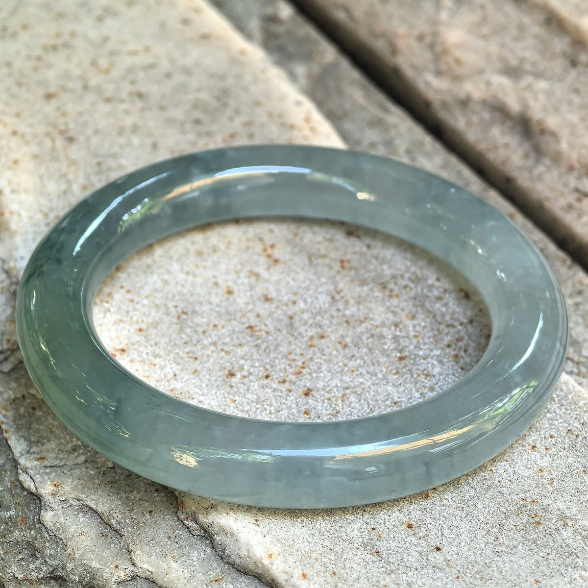 Angled view of the light green round-bar jadeite bangle on stone, highlighting its rounded profile, subtle blue undertone, and gel-like luster.