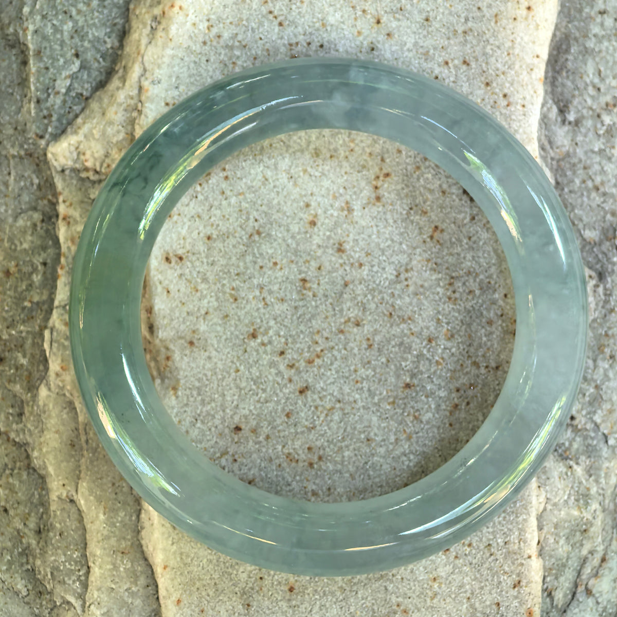 Overhead view of the light green round-bar jadeite bangle resting on stone, showing its circular outline and polished inner and outer surfaces.