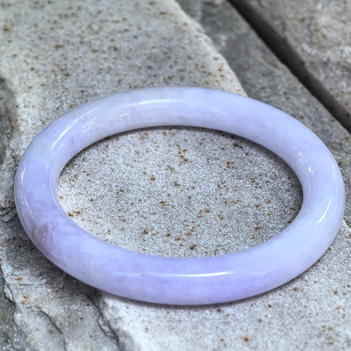Angled close-up of a lavender round-bar jadeite bangle on stone, showing its polished surface and a localized patch of mottled cotton-like inclusions.