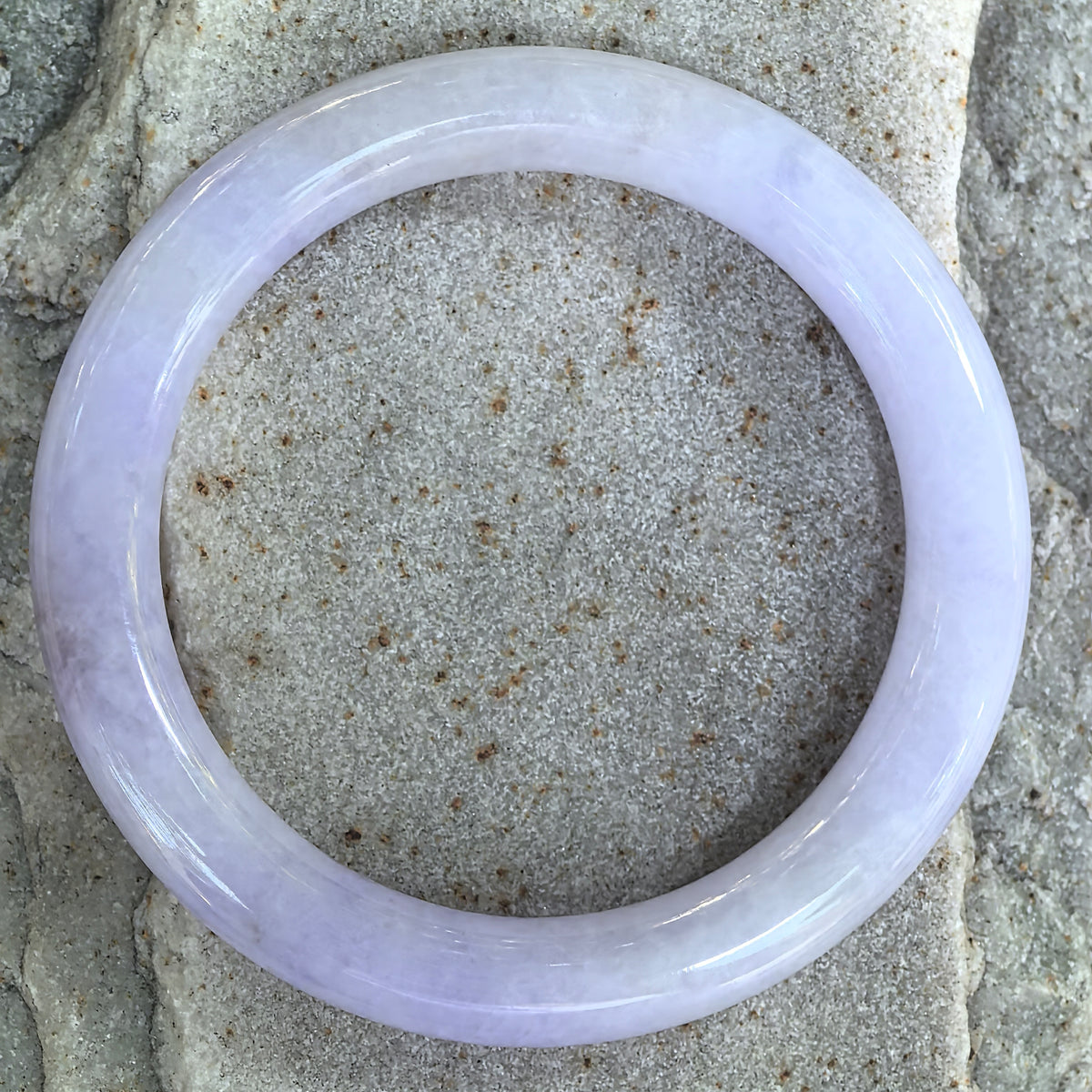 Overhead view of a lavender round-bar jadeite bangle on stone, showing the circular shape and variations in its natural texture.