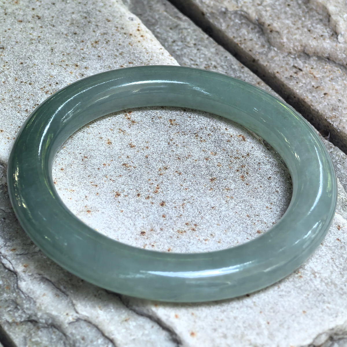 Angled view of the green round-bar jadeite bangle resting on stone, showing its rounded profile and polished inner and outer surfaces.