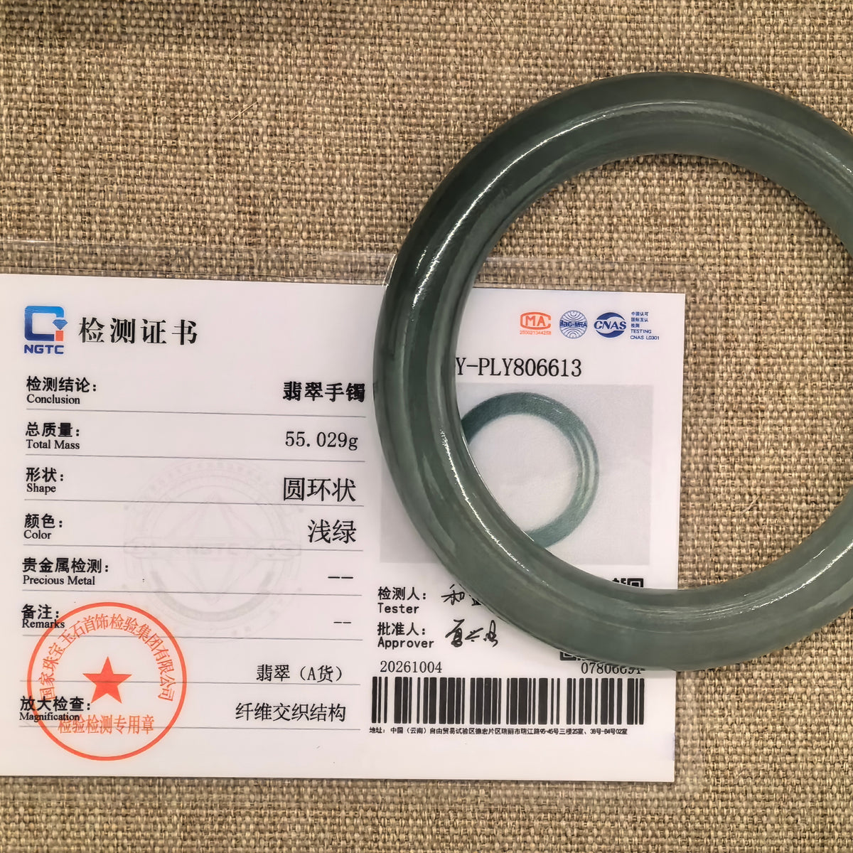 Green round-bar jadeite bangle displayed with its matching NGTC certificate numbered Y-PLY806613 on a woven background.