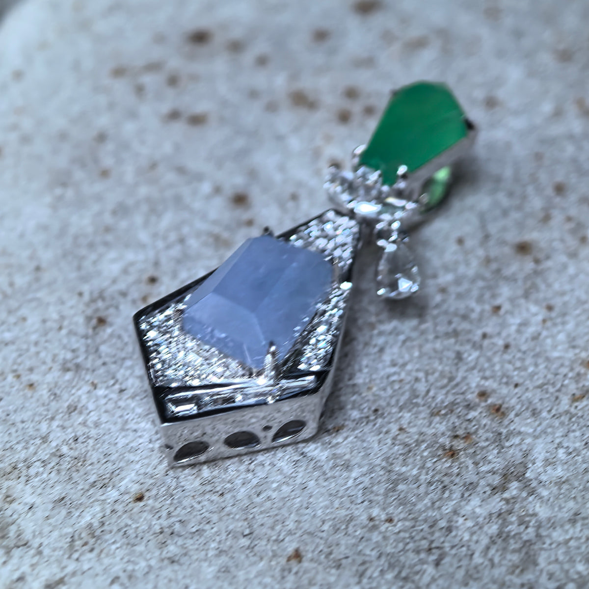 Low angled view of a lavender and green jadeite geometric kite-shaped pendant on stone, showing the raised setting and side profile.