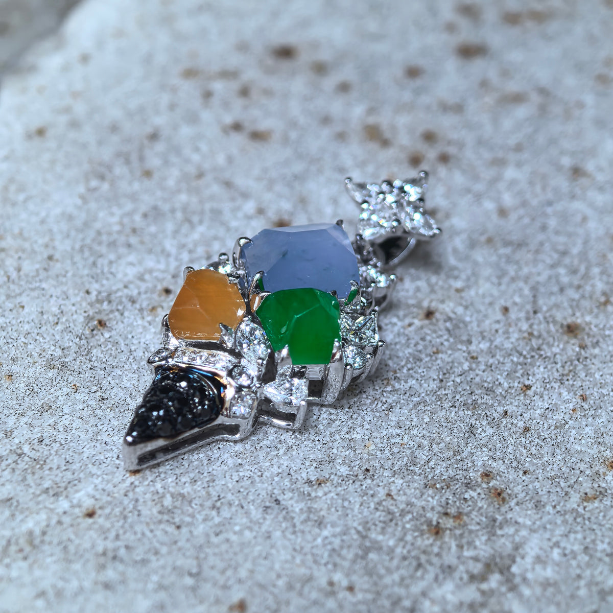 Low angled close-up of a multicolor jadeite ice cream cone pendant on stone, showing the jadeite contours, 18K gold setting and black cone accents.