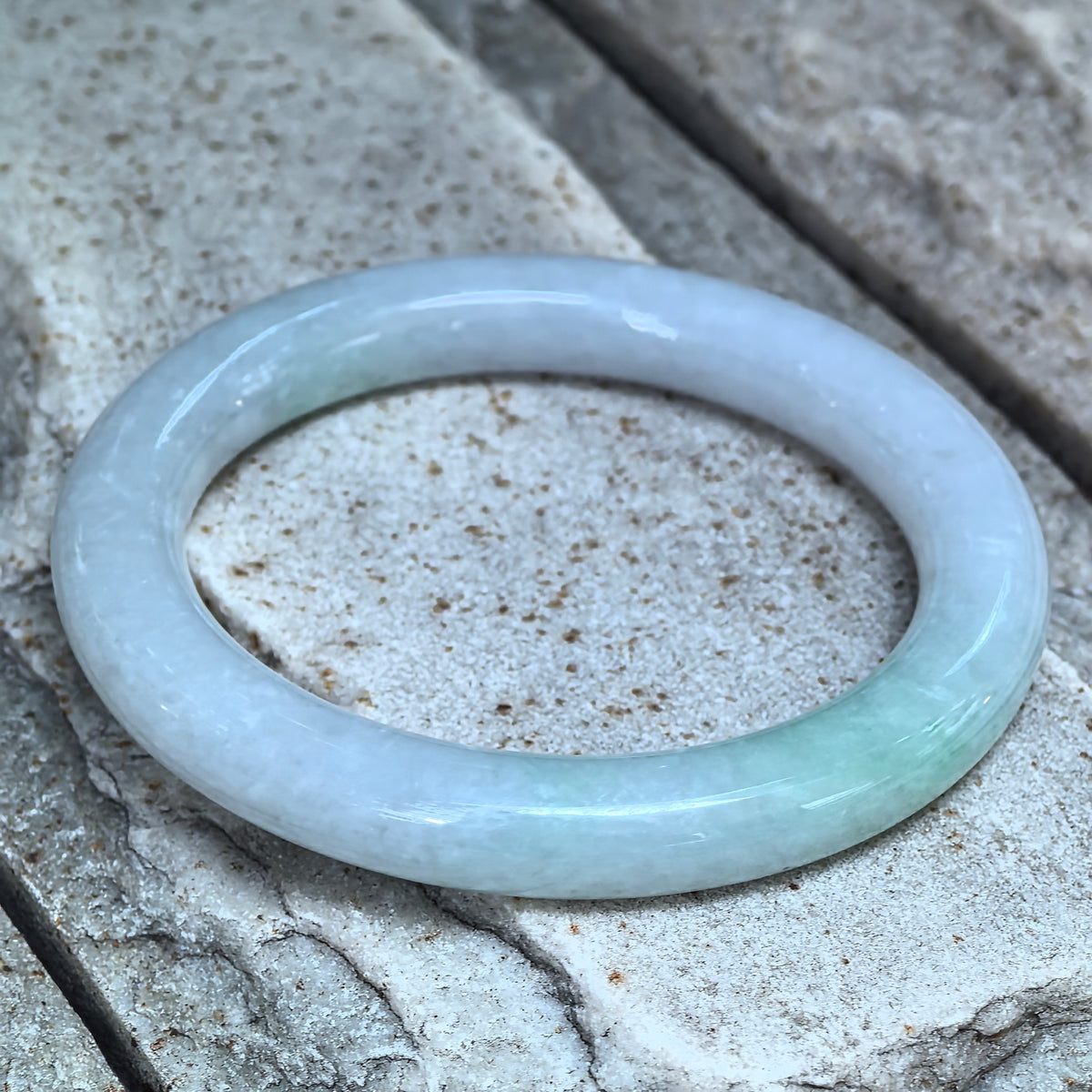 Angled view of a pale green and lavender round-bar jadeite bangle resting on stone, showing its rounded inner and outer surfaces.