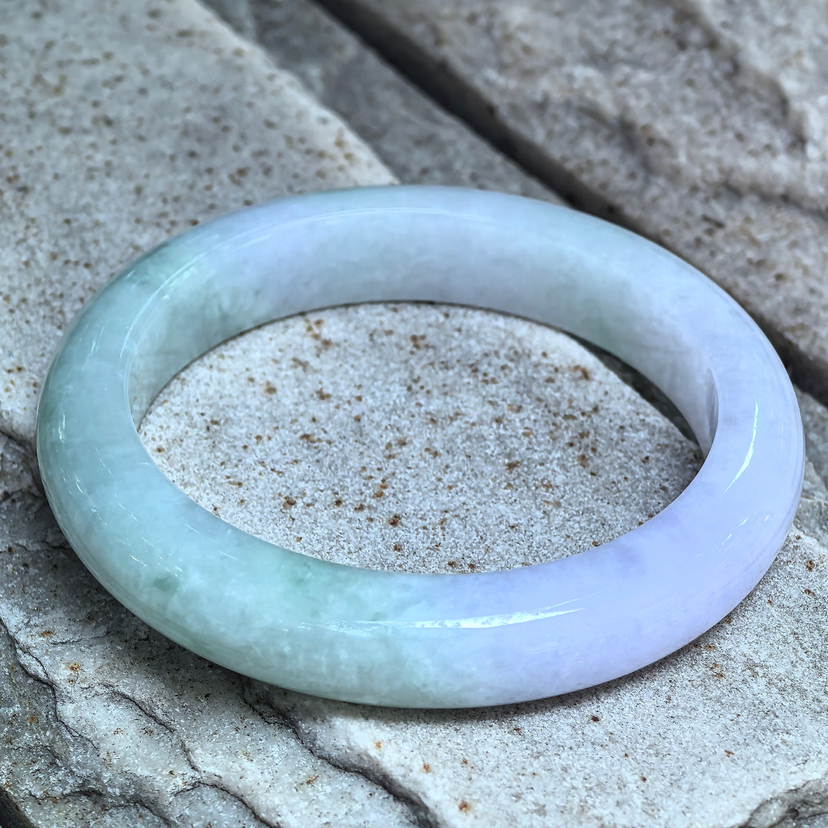 Angled view of a pale green and lavender D-shape jadeite bangle on stone, showing its rounded exterior and flat inner surface.
