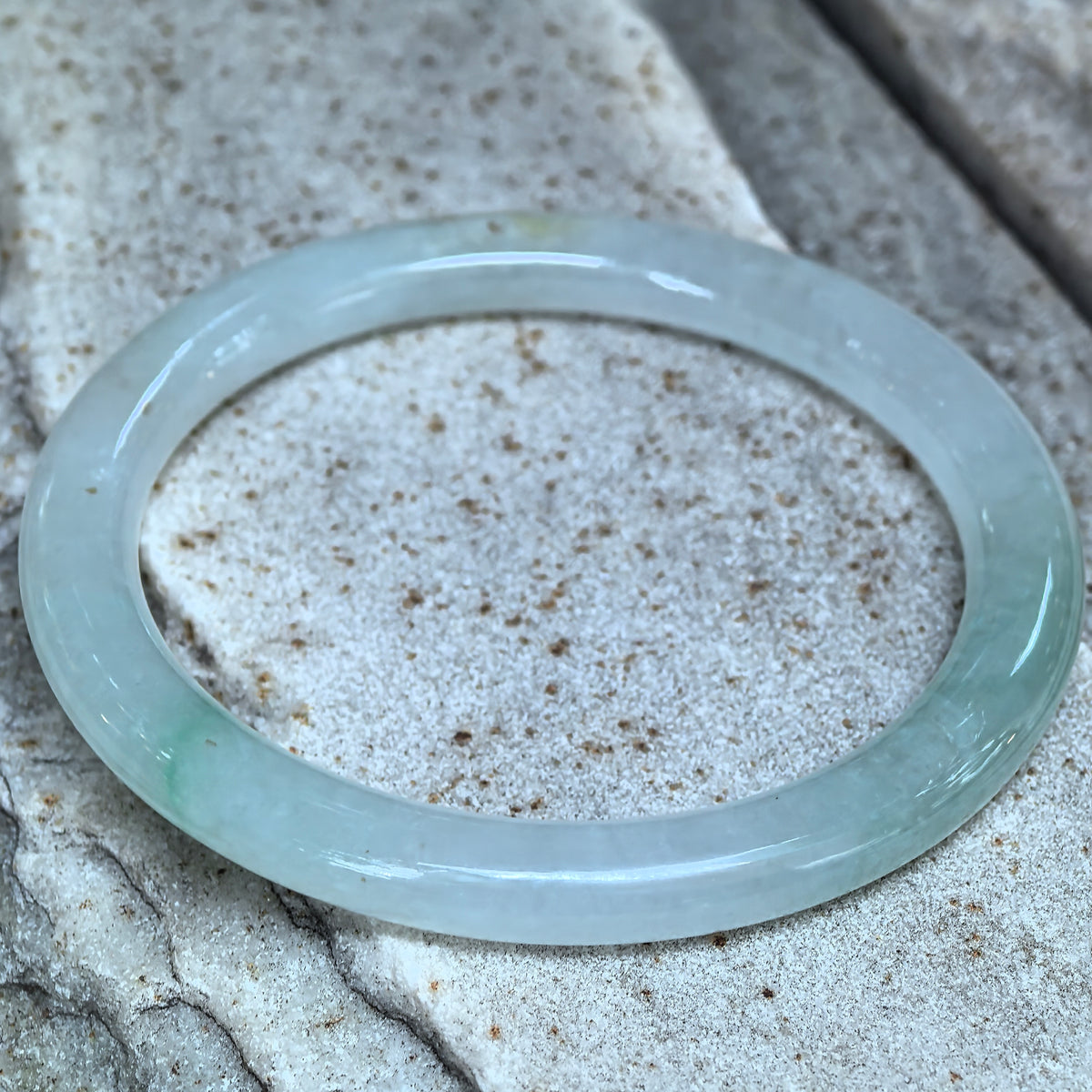 Angled close-up of a light green round-bar jadeite bangle on stone, showing a few natural golden-brown flecks and its polished finish.