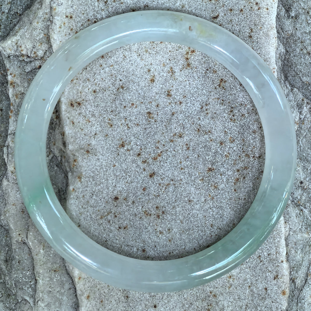 Overhead view of a slender light green round-bar jadeite bangle on stone, showing scattered golden-brown flecks around the band.