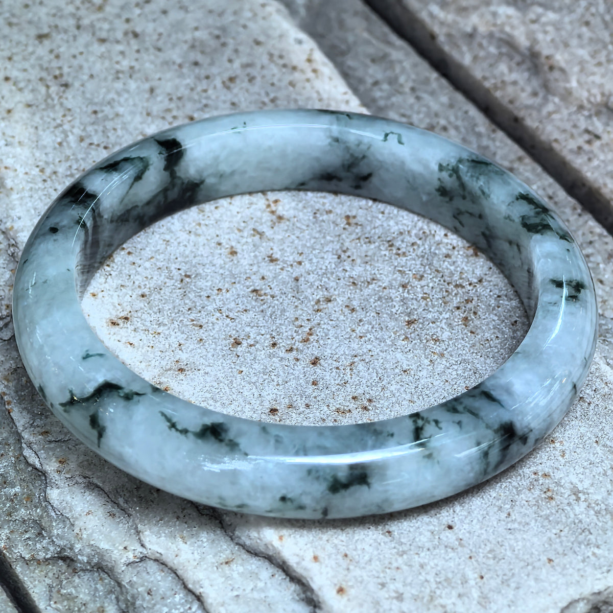 Angled close-up of a D-shape jadeite bangle on stone, showing deep green floating patterns and the flat inner surface.