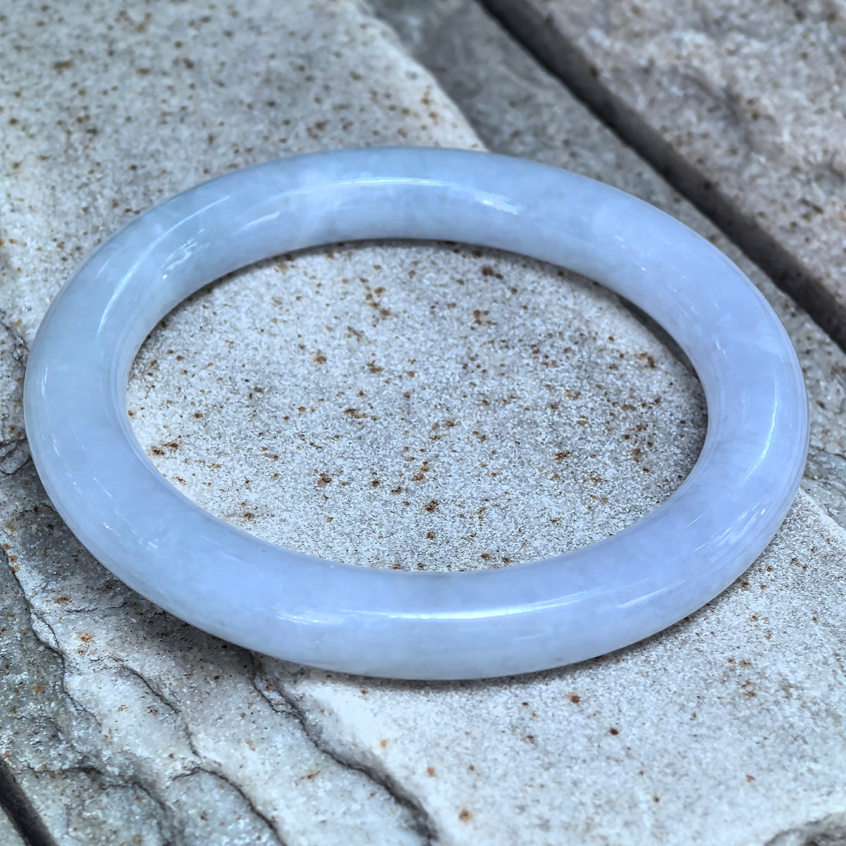Angled view of a glutinous pale lavender round-bar jadeite bangle resting on stone, showing its rounded contours and polished luster.